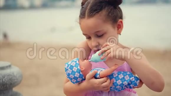 可爱的小女孩用玩具瓶子喂她最喜欢的娃娃幼儿在海边的公园里玩娃娃开心快乐视频的预览图