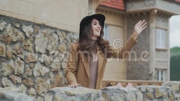 一位戴着帽子的漂亮年轻女子站在一个石质阳台上挥舞着她的手和一个陌生人说话视频的预览图