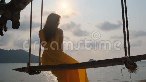 年轻漂亮的女孩穿着黄色的裙子在秋千上海边海洋视频的预览图