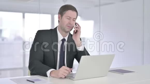 中年商人在智能电话上交谈在笔记本电脑上工作视频的预览图