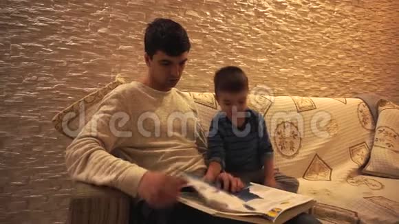 爸爸穿着白色的毛衣黑发小儿子穿着蓝色的条纹看着百科全书坐在一个柔软的衣服上视频的预览图
