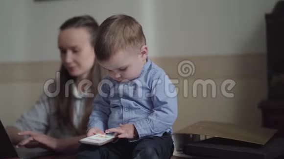 一位年轻的母亲一位在办公室工作的女商人带着笔记本电脑一个可爱的小男孩坐在桌子上视频的预览图