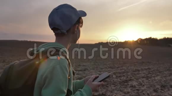 带背包的有趣男孩在日落时走在户外使用电话听音乐旅行的概念视频的预览图