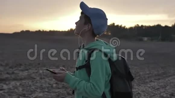 带背包的有趣男孩在日落时走在户外使用电话听音乐旅行的概念视频的预览图