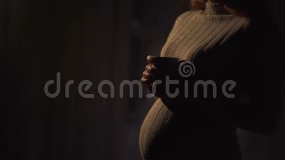不明身份的怀孕女孩站在温暖的毛衣在黑暗的房间里喝热茶女士在等视频的预览图