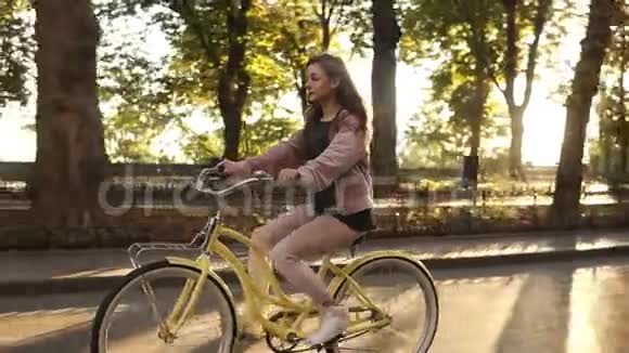 侧景漂亮的长发女孩骑自行车在街道或林荫大道在夏季城市公园穿着粉色衣服视频的预览图