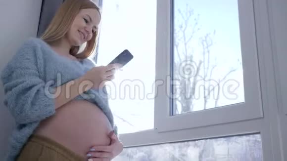 怀孕快乐的女孩抚摸她赤裸的大肚子在大窗户附近的背光中使用手机视频的预览图