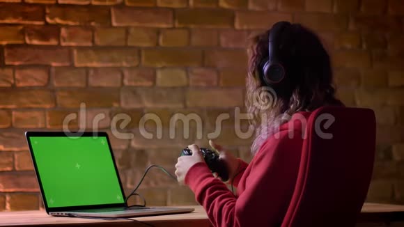 穿红色连帽衫和戴耳机的年轻博主用笔记本电脑和布里克肯墙上的操纵杆玩电子游戏的后视图视频的预览图