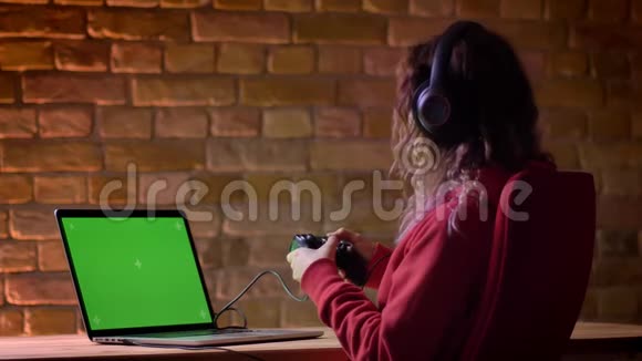 耳机中的年轻博客作者使用笔记本电脑和布里克肯墙上的操纵杆积极玩电子游戏的后视镜视频的预览图