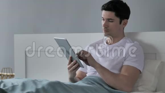 年轻人在床上的平板上浏览互联网视频的预览图
