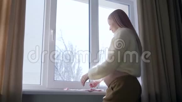 未来的妈妈手里拿着孩子的衣服站在家里背光的窗户附近视频的预览图