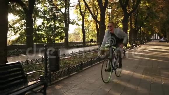 穿着深色牛仔裤蓝色衬衫和太阳镜的年轻人独自在城市公园骑自行车停在长凳附近视频的预览图