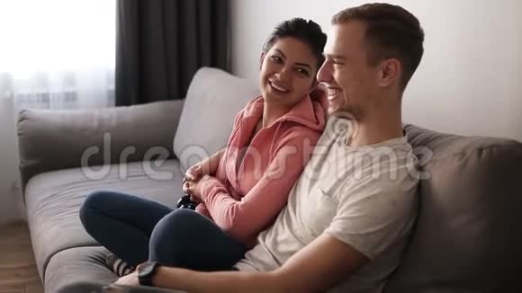 年轻夫妇在明亮的客厅里的灰色沙发上看电视令人兴奋的一对夫妇对电视节目感兴趣侧面视频的预览图