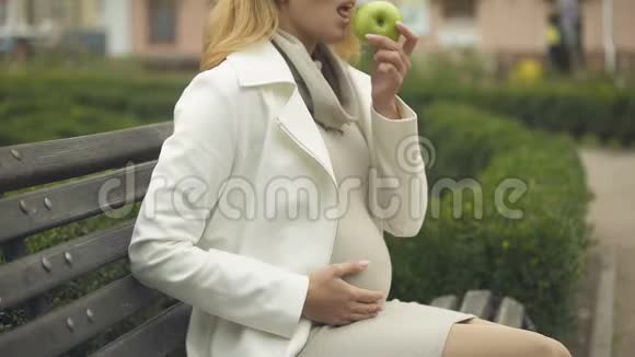 金发孕妇吃绿苹果在长凳上放松产妇幸福视频的预览图