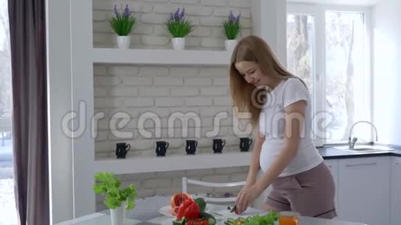 怀孕和健康的营养美丽的女孩切蔬菜吃西红柿在厨房里抱着她的大肚子视频的预览图