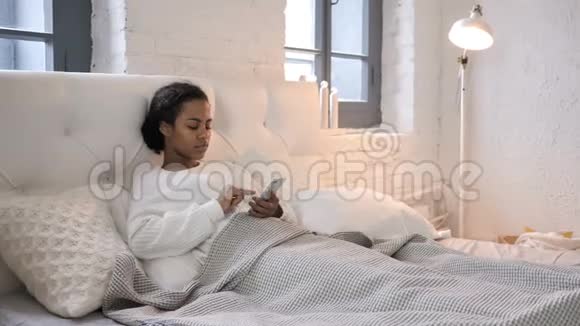 年轻的非洲女孩在床上放松时使用智能手机视频的预览图