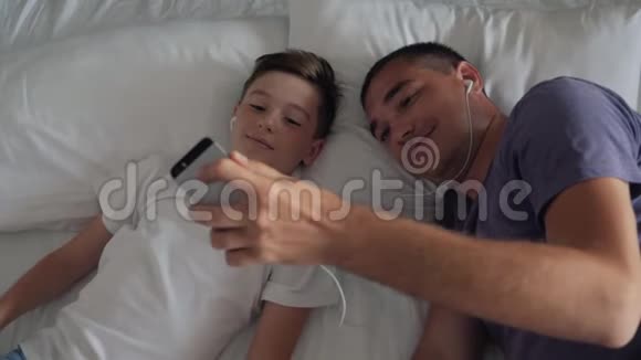 一个幸福的家庭父子俩躺在床上听着耳机里智能手机上的音乐视频的预览图