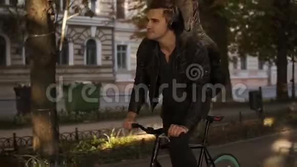 布鲁内特英俊的年轻人穿着黑色便服戴着耳机骑着自行车在铺好的城市公园里吉他在里面视频的预览图