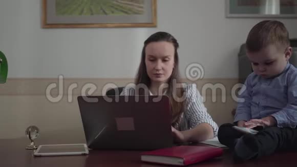 肖像美丽的母亲女商人在办公室桌上工作手提电脑和可爱的小男孩坐在桌子上视频的预览图