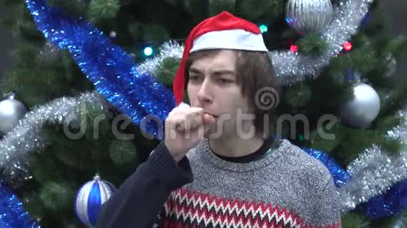英俊的年轻人戴着圣诞帽在圣诞树背景上咳嗽视频的预览图