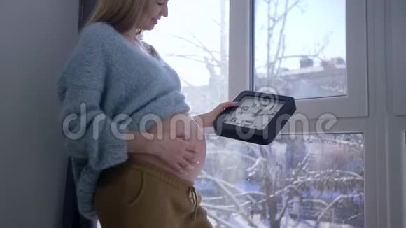 怀孕的女孩手里拿着时钟站在一扇大窗户附近抚摸着她赤裸的大肚子视频的预览图