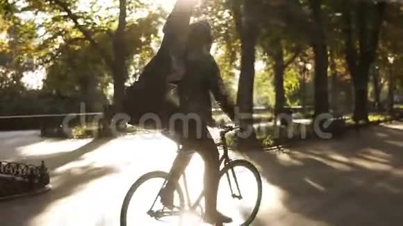 一个穿着黑色便服的年轻人戴着黑色吉他的耳机骑着自行车骑在铺成的城市公园旁视频的预览图