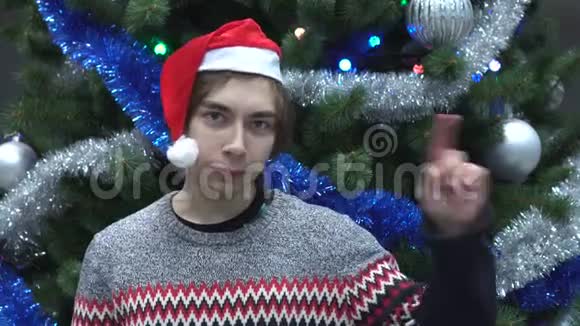 戴着圣诞帽和挥舞手指的英俊青年在圣诞树背景上拒绝视频的预览图