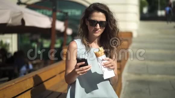 美丽的年轻女子吃羊角面包和看电话附近的户外咖啡馆视频的预览图