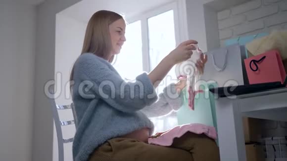 美丽的怀孕女孩期待孩子看着粉红色的婴儿衣服抚摸着赤裸的肚子坐在桌子旁视频的预览图