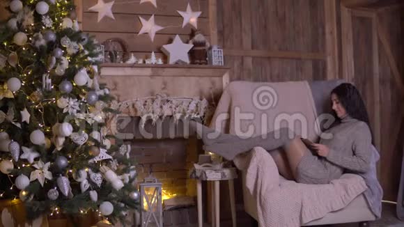 一个留着深色长发的年轻女孩正坐在圣诞树附近的扶手椅上看书4K视频的预览图