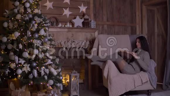 一个留着长发的女孩打开一个装有礼物的盒子并在圣诞节的时候坐在圣诞树附近的椅子上高兴视频的预览图