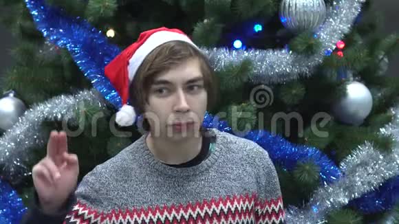 英俊的年轻人戴着圣诞帽偷听圣诞树的背景视频的预览图
