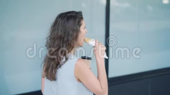 年轻的金发女人吃羊角面包喝咖啡路过大楼视频的预览图