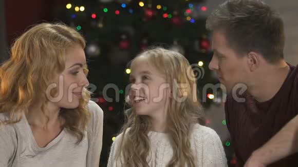 快乐的父母亲吻他们可爱的女儿的脸颊坐在圣诞树下视频的预览图