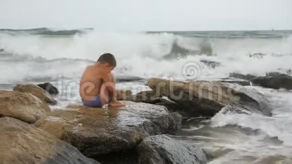 孩子在海边冲浪男孩坐在一块大石头上享受海浪的声音视频的预览图