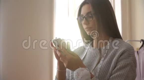 带眼镜的年轻俄罗斯女裁缝对着窗户看电话从阳光照耀的地方视频的预览图