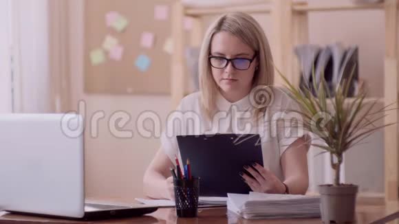 金发女孩白种人的外表与眼镜和一件白色衬衫在办公室工作与文件和笔记本视频的预览图