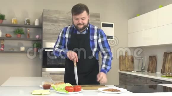 一个穿着围裙的男厨师在厨房里切一个西红柿桌子上摆着汉堡的配料慢悠悠视频的预览图