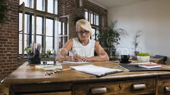 一位有魅力的成年女士坐在办公桌后的办公室里工作时会在笔记本上记录一些东西视频的预览图