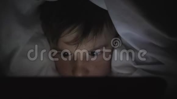 一个快乐的男孩躺在毯子下的床上在黑暗中玩平板电脑孩子的脸被视频的预览图
