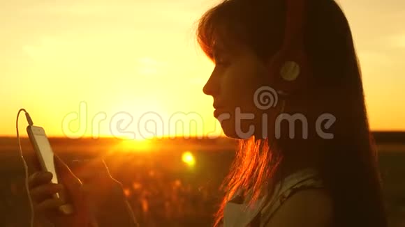 女孩旅行者在智能手机上听音乐在美丽的春天日落的光线下带耳机的年轻女孩视频的预览图
