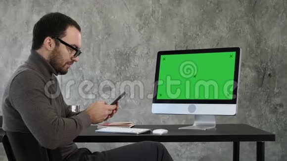 商人看着他的智能手机短信坐在电脑屏幕附近绿色屏幕模拟显示视频的预览图
