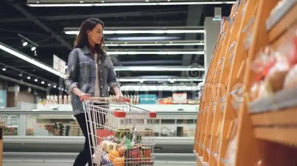 微笑的女孩正在超市买新鲜面包闻一闻然后把它和其他产品一起放进手推车里购物视频的预览图