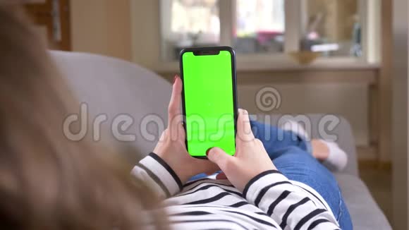 特写后景拍摄女性在手机上观看视频绿色屏幕躺在舒适的室内沙发上视频的预览图