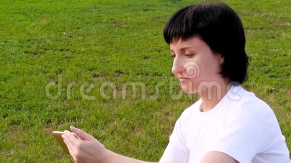 一个黑发女孩坐在绿草上用智能手机一位年轻女性在社交网络中发送信息视频的预览图