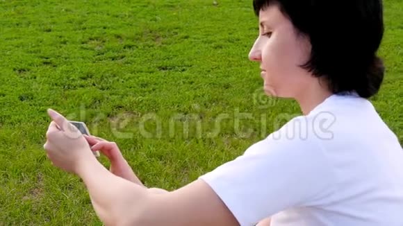 布鲁内特女孩坐在公园的绿草上使用智能手机一个年轻女人在社交场合发信息视频的预览图