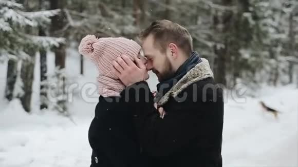 年轻漂亮的白种人夫妇在冬天的森林里在雪地里接吻一对玩得开心男人把帽子拉到她身上视频的预览图
