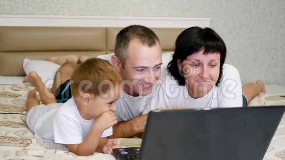 幸福的一家人妈妈爸爸和他们的小儿子正在笔记本电脑上看电影躺在床上表现出情绪视频的预览图