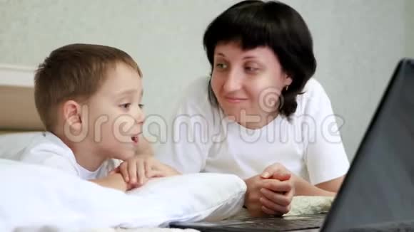 幸福的母亲和她的孩子躺在床上微笑着看着笔记本电脑屏幕视频的预览图