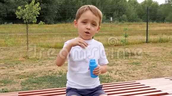 一个快乐的孩子正坐在操场的长凳上缓慢地吹着泡泡视频的预览图